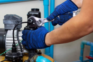 Close-up de técnico realizando manutenção com luvas azuis usando chave inglesa na manutenção de motobomba doméstica, com tubulação e elétrica.